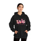 female model wearing hope + joy collective black hoodie sweatshirt with "Tis the Season" in cursive font below a graphic image of seasonal coffee and cocoa drinks accented in pink with a pink Christmas tree topped with pink bow