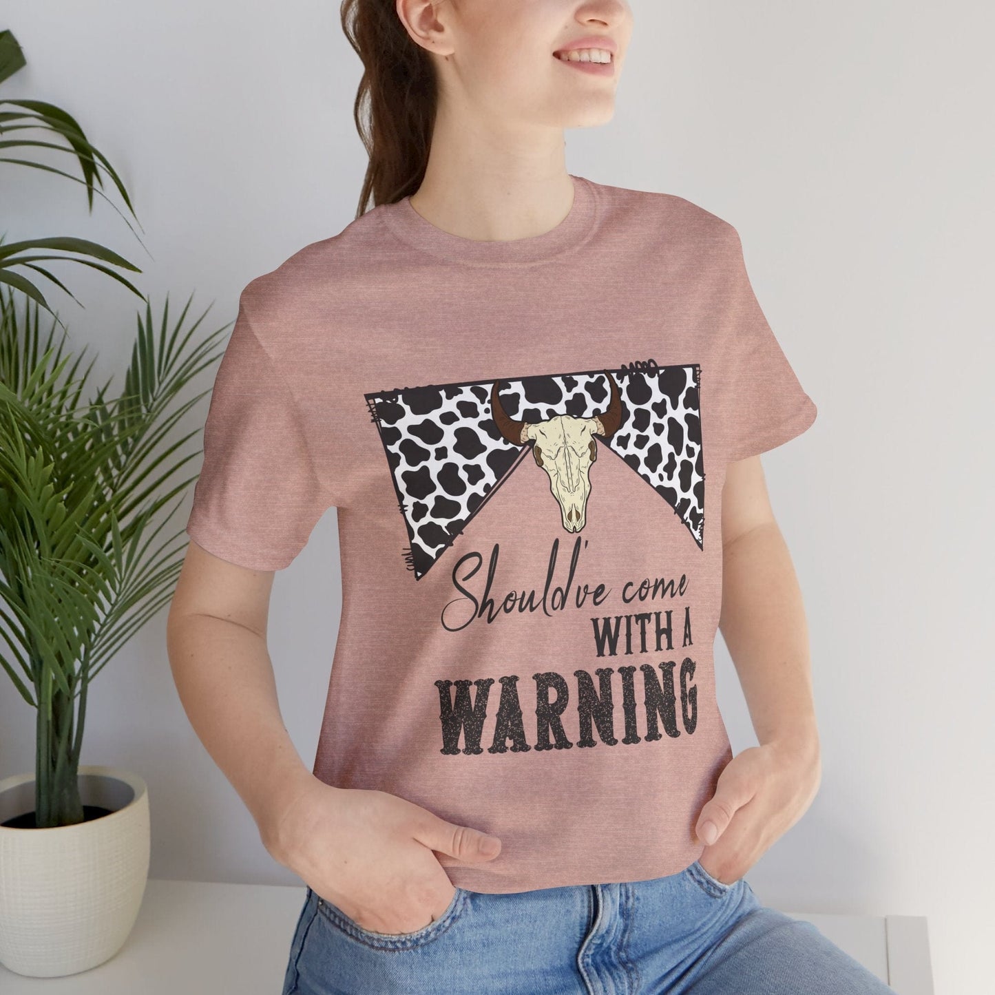 female model in jeans wearing hope + joy collective heather prism peach short sleeve t-shirt with a graphic image of a leopard banner and cow skull in the middle that says "Should've Come with a Warning" in western font