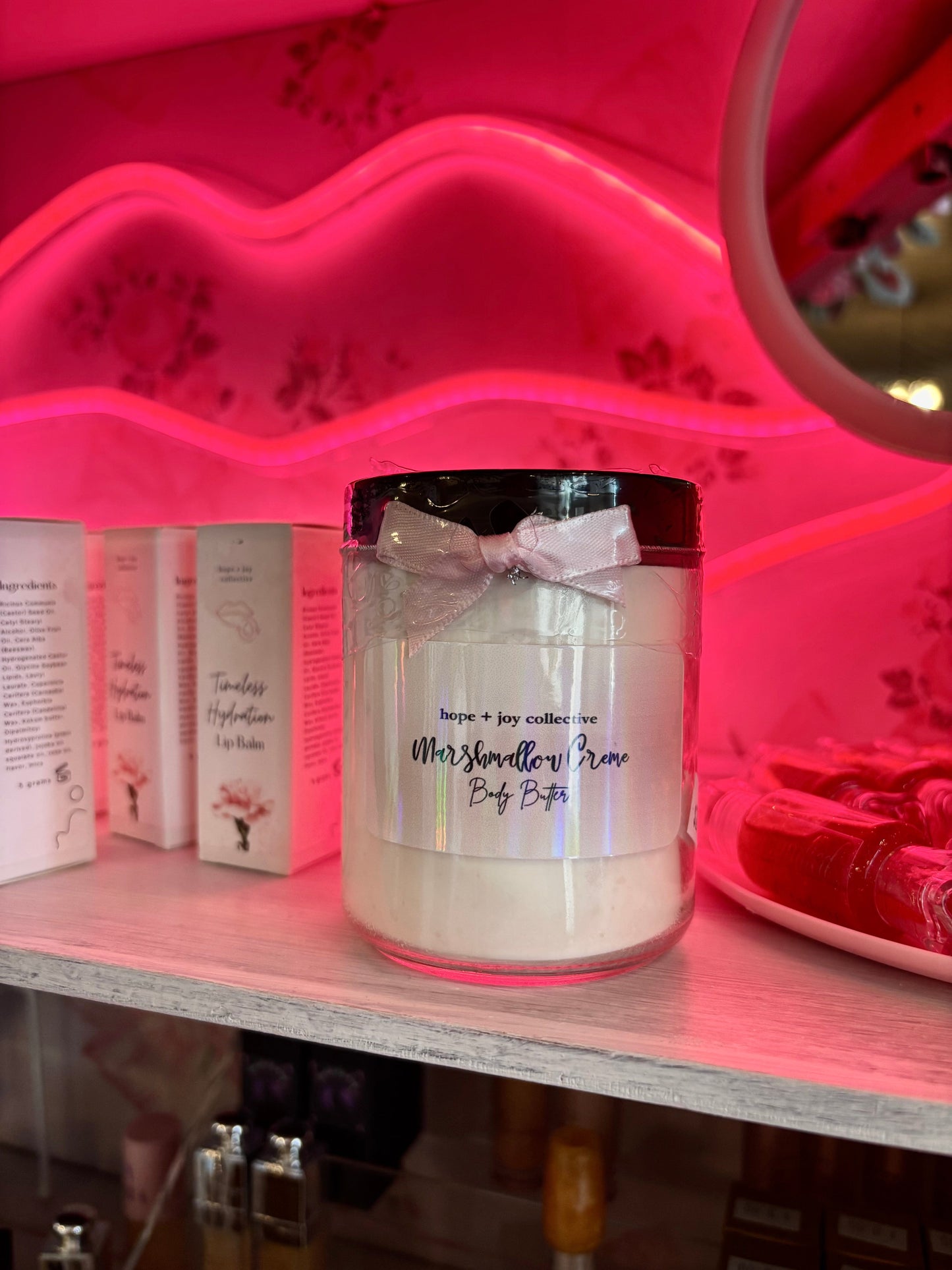 hope + joy collective Marshmallow Creme Body Butter in glass jar with small pink ribbon bow on shelf with neon pink lip light in background