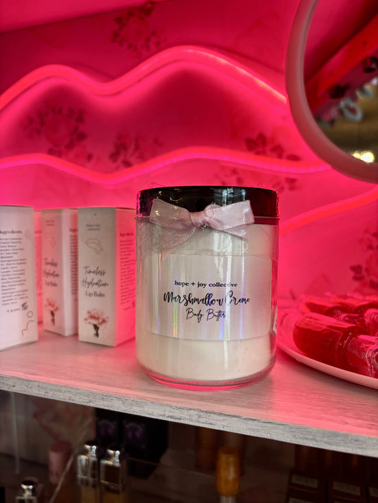 hope + joy collective Marshmallow Creme Body Butter in glass jar with small pink ribbon bow on shelf with neon pink lip light in background