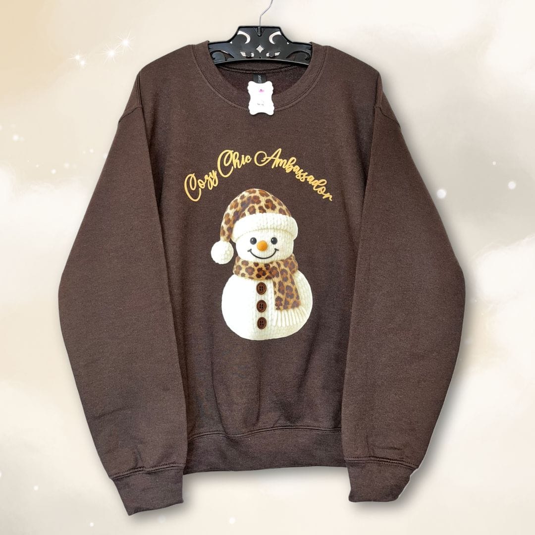hope + joy collective Cozy Chic Ambassador Snowman Crewneck brown sweatshirt with realistic knit-look image of snowman wearing and leopard print hat and scarf and "Cozy Chic Ambassador" in swirly puff textured font arching over the snowman.