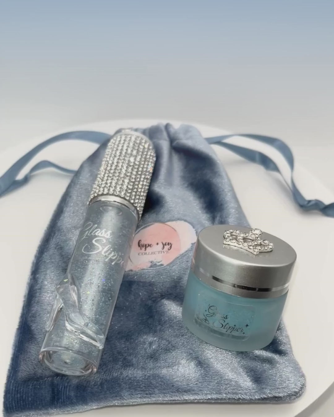 video of hope and joy collective Glass Slipper Lip Gloss and Lip Scrub Set on blue velvet pouch with HJC pink round logo rotating on white base. Lip gloss tube has silver rhinestone cap and clear molded resin women's high heel shoe on bottom front.  Frosted glass lip scrub jar has blue contents showing through with matte silver lid and rhinestone crown on top.