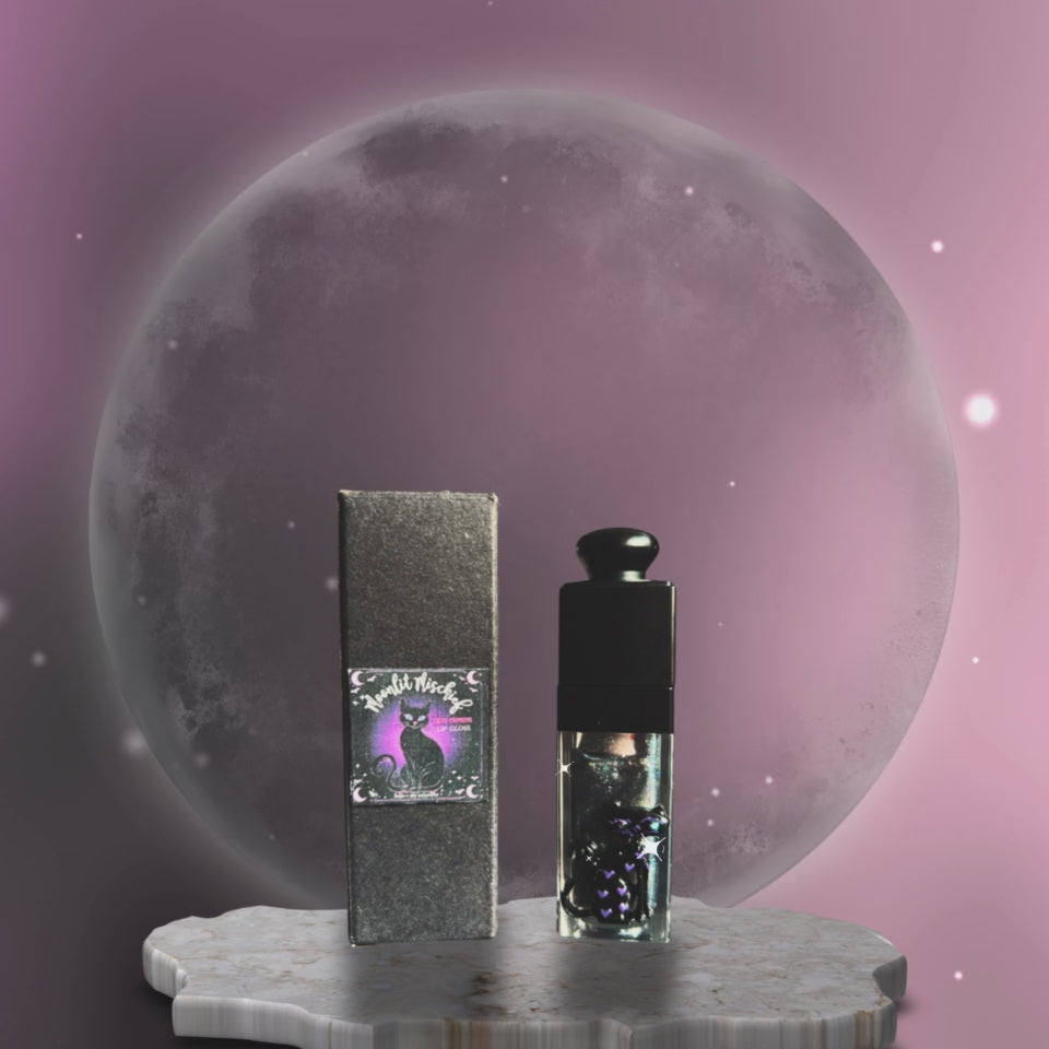 hope and joy collective video of  Moonlit Mischief Lip Gloss tube, black cap and 3D cat on front with rhinestone eyes and labeled box packaging standing on a gray irregular shaped marble stand with a glowing moon in the background and a dark and light purple moving sky with glistening starts.